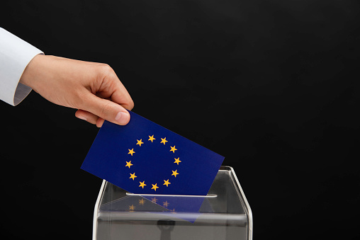 Human hand is inserting European Union flag into ballot box in front of black background. Representing elections in  European Union.