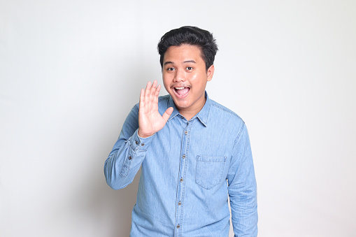 Portrait of excited Asian man in blue shirt whispering malicious talk conversation, hand on mouth telling secret rumor. Isolated image on white background