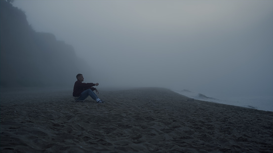 Sad man looking sea landscape in fog. Wide shot serious guy sitting on sandy beach. Male person relaxing on ocean shore in autumn morning. Calm man enjoying nature. Mysterious natural scene in mist
