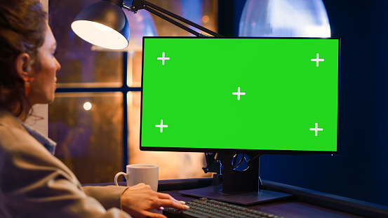Young entrepreneur looking at greenscreen display on monitor, checking computer with chromakey blank template. Startup manager working after hours with isolated copyspace screen.