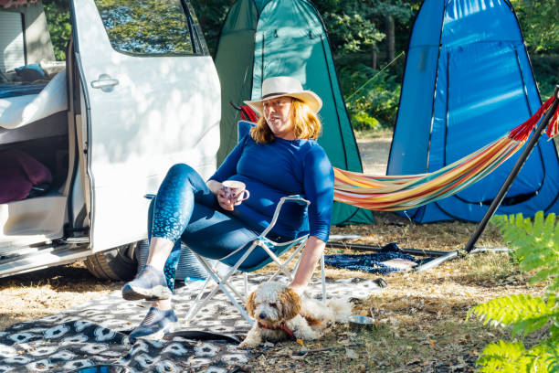 erwachsene frau entspannt sich, trinkt tee im reisestuhl und streichelt ihren cockapoo-welpen in der nähe von wohnmobil und zelten. frau lebt auf einem wohnmobil mit tier und bereist die welt. bester freund. - camping mobile home vacations tent stock-fotos und bilder