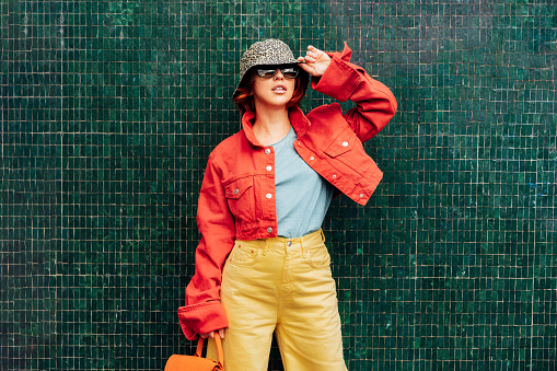Hipster young woman in bright clothes, sun glasses, backpack bag and bucket hat posing on the green tile wall background. Urban city street fashion. Fashion blogger. Selective focus. Copy space,