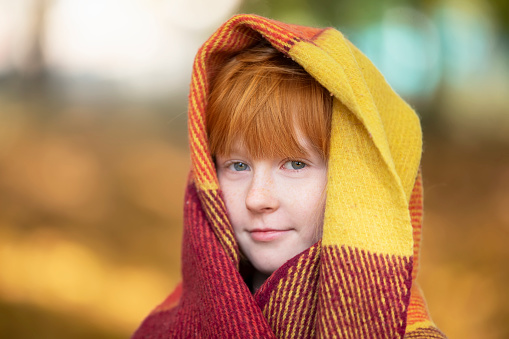 A red-haired girl wrapped herself in a woolen blanket in an autumn park. Child in autumn.