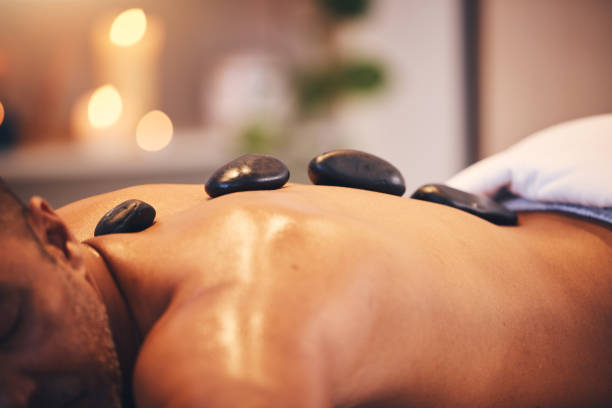 masaje, piedras calientes y relajarse con un hombre en un spa, acostado en una mesa o cama para terapia física en un resort de lujo. wellness, rock y zen con un cliente masculino relajándose en un centro de salud para descansar - lastone therapy men spa treatment male fotografías e imágenes de stock