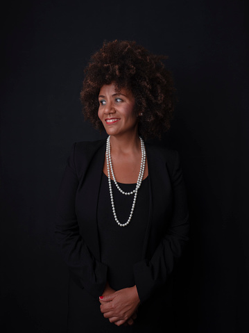 elegant woman dressed in a dark colored suit and jacket, black woman with afro hair, looking to the side with a smile. wearing a pearl necklace. dark background.