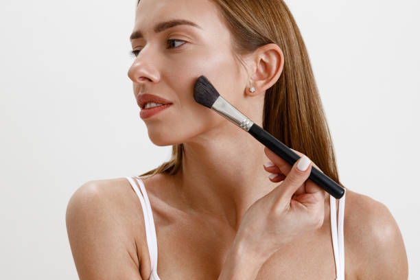 istock Pretty woman using cosmetic brush to apply decorative makeup on her face on studio background 1466011485