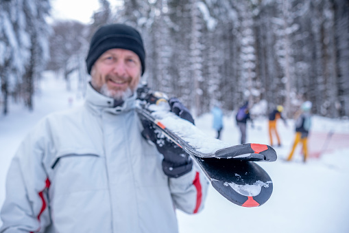 A cheerful man in a red-white ski outfit, black hat, and gloves with a ski on his shoulder is looking at the camera. The focus is on the tip of the skis. The face is out of focus.