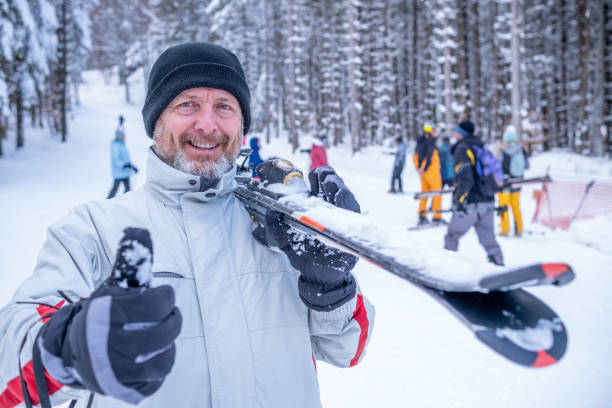 Cheerful man in a ski outfit and skis on his shoulder on a ski slope. A cheerful man in a red-white ski outfit, black hat, and black gloves with a ski on his shoulder is looking at the camera. The man has a thumbs up. thumbs up for skiing stock pictures, royalty-free photos & images