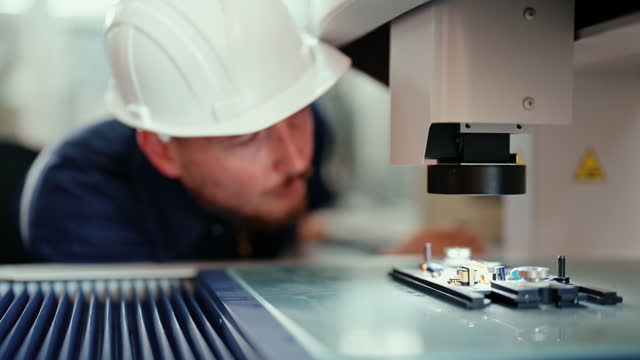 Caucasian production engineer in safety wear is checking for defects on electronics board by using a microscope.