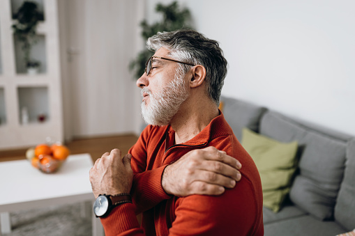 Side view mature Caucasian man sitting on living room sofa holding his painful shoulder