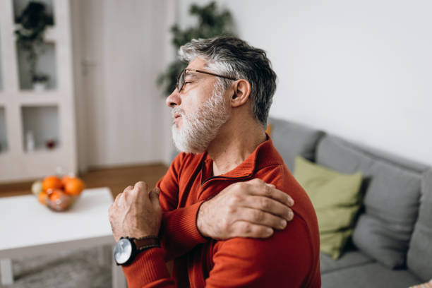 Mature man having problems with joints Side view mature Caucasian man sitting on living room sofa holding his painful shoulder joint pain stock pictures, royalty-free photos & images