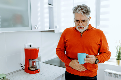 Poor mature Caucasian man standing in his kitchen drinking coffee suffering from stomachace