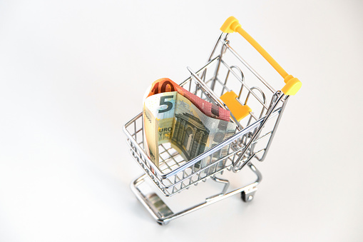 Supermarket shopping cart\nwith euro banknotes inside on white background. Conceptual image about purchasing power, family expenses, essential goods, consumer society. Copy space.