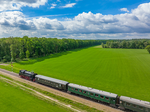 Steam train with smoke from the locomotive driving in the countryside with smoke coming from the chimney seen from above. The black and red locomotive is pulling passenger railroad car with tourists. Aerial drone photo.