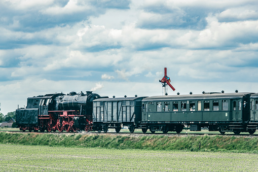 Steam train with smoke from the locomotive driving in the countryside with smoke coming from the chimney. The black and red locomotive is pulling passenger railroad car with tourists.