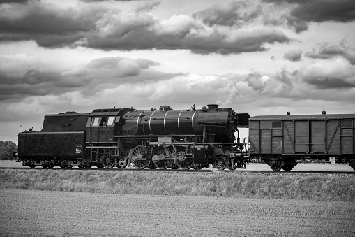 Steam train with smoke from the locomotive driving in the countryside with smoke coming from the chimney. The black and red locomotive is pulling passenger railroad car with tourists. Black and white photograph.