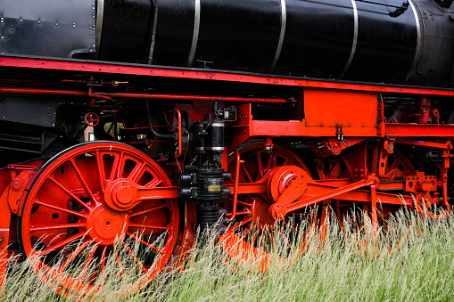 Steam train wheels driving in the countryside with smoke coming from the chimney. The black and red locomotive is pulling passenger railroad car with tourists.