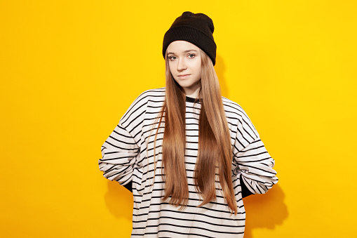 Studio portrait of a modern young white woman with long brown hair in a striped sweatshirt, in a black knitted hat, against a yellow background