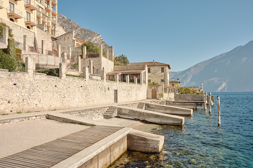 Wide-angle view of typical multi-storey stonework buildings overlooking a little harbour in Limone sul Garda, still serving their original function of enclosing gardens, lemon tree orchards and olive groves (locally known as limonaie and oliveti respectively). The hazy cliffs and slopes of the mountain range of Monte Baldo can be seen on the background. A perfectly clear sky, the golden oblique light of a late afternoon, long shadows, low contrast. Palette based on pastel tones, ranging from creamy whites to bright yellows and pale cyans, with green accents. High level of detail, natural rendition, realistic feel. Developed from RAW.