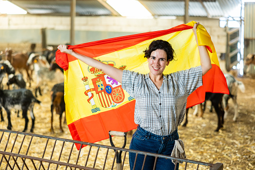 Joyful young Latin female traveler waving Spanish flag inside of livestock goat farmhouse