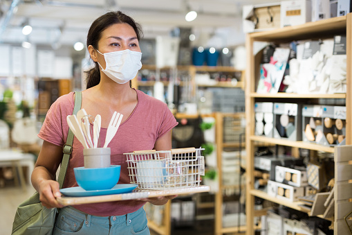 Female customer in protective mask holding purchases and walking in a household goods store