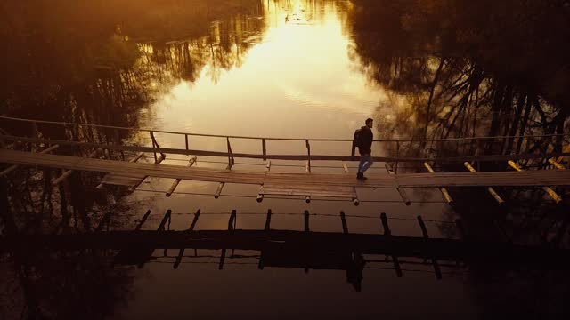 Aerial man walking along wooden suspension bridge over river in evening golden hour, trekking in autumn forest, swinging footbridge