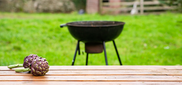 Artichoke on a wooden table outdoors on a blurred grill background. Preparing to cook green grill. Banner, place for text.