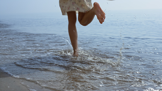 Bare woman legs jumping on sea waves close up. Unknown young lady wearing cute dress have fun walking on seashore. Slim wet girl feet splashing in ocean water summer weekend. Happiness concept.