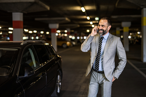 Portrait of happy mid adult businessman talking on his phone while standing in public garage next to his car.