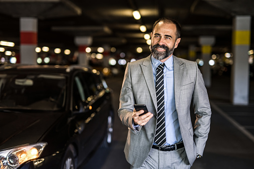 Mid adult businessman wearing a suit is standing next to his car and using his cell phone at public garage.