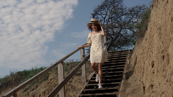 Energetic smiling woman hurrying downstairs sand beach holding straw hat on head. Cute happy girl in flowery dress running on summer seashore. Attractive curly lady have fun joy on beautiful nature.