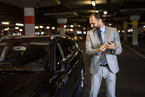 Mid adult businessman wearing a suit is leaving his car and using his cell phone at public garage. He is looking at his beautiful car.