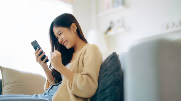 Happy young asian woman relax on comfortable couch at home texting messaging on smartphone, smiling girl use cellphone, chatting online message, shopping online from home stock photo