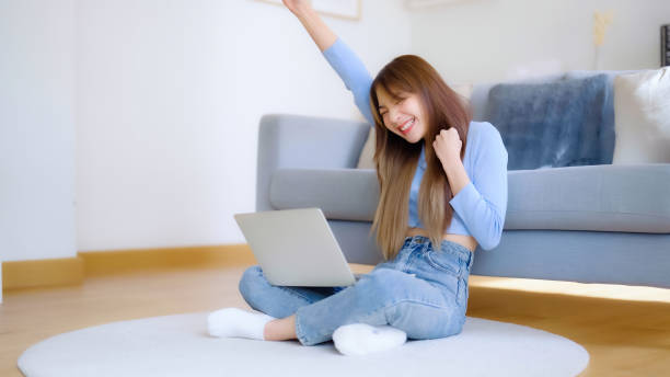 Young asian woman in good spirits working on laptop at home while sitting on the floor close to the couch. Excited female winner celebrating success while glancing at notebook stock photo