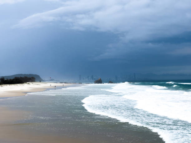 Horizontal landscape of storm clouds over the distant Surfer’s Paradise skyline and ocean waves breaking onto the sand beach at Currumbin, Queensland.