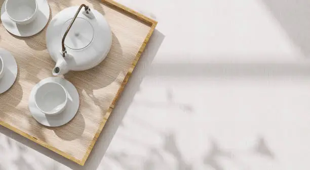 Photo of Top view of modern, minimal, beautiful white ceramic teapot, teacup with saucer on brown straw mat wooden tray on cream tablecloth in sunlight and leaf shadow