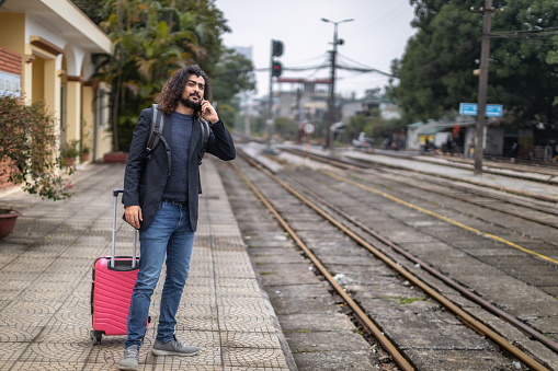 Young man with long curly hair and beard waiting for a train on a rail station platform, wearing a suit jacket, carrying a backpack and pink suitcase, at the Gia Lam train station, Long Bien, Hanoi, Vietnam