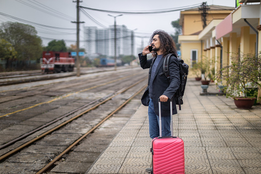Young man with long curly hair and beard waiting for a train on a rail station platform, wearing a suit jacket, carrying a backpack and pink suitcase, at the Gia Lam train station, Long Bien, Hanoi, Vietnam
