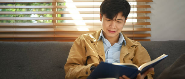 istock Pleased asian man sitting on comfortable sofa and reading interesting book, enjoying leisure weekend time at home. 1466264138
