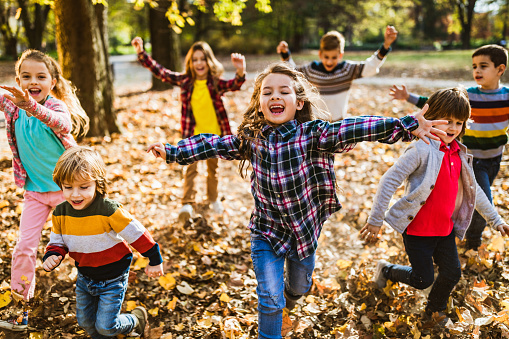 A group of multiethnic school aged children are scooping the leaves up in their arms and throwing them in the air.  They are each dressed casually in warm fall attire and laughing as the colorful leaves fall to the ground overhead.