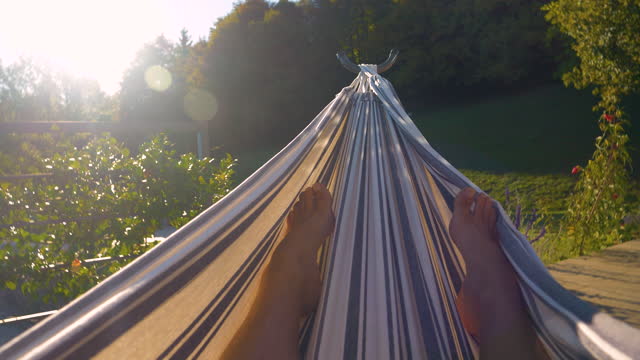 POV, LENS FLARE: Young person relaxing in swinging hammock in summer garden