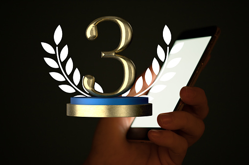 A 3D model of a 3rd place trophy near a lit screen of a smartphone in hand on a dark background