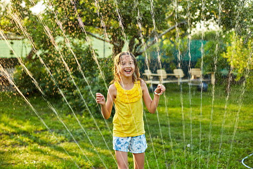 Happy kid girl playing with garden sprinkler run and jump, summer outdoor water fun in the backyard at home, splash on sunny day.