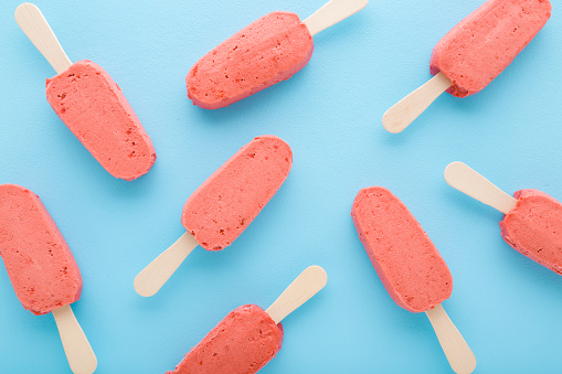 Red strawberry ice cream with sticks on light blue table background. Pastel color. Closeup. Cold sweet snack in summer. Food pattern. Top down view.