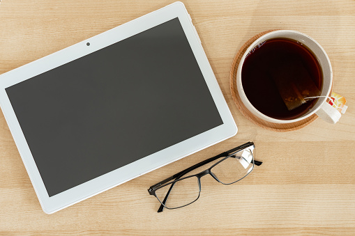 A top view of a tablet, eyeglasses, and a teacup on a wooden office desk