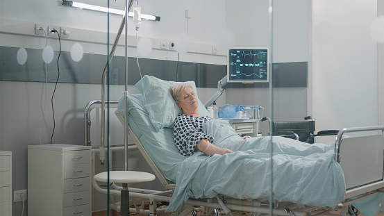 Medical assistant looking at heart rate monitor while patient sleeping in hospital ward bed. Nurse checking up on retired woman with nasal oxygen tube for healthcare and recovery.