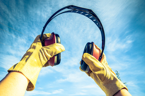 A male worker's hands in yellow gloves holding ears muffler under a wispy sky