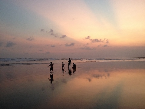 Silhouette of a small family playing in Parangtritis beach at sunset with horizon and mirror reflection of sky, clouds and shadows on sand for tourism travel concept and background.