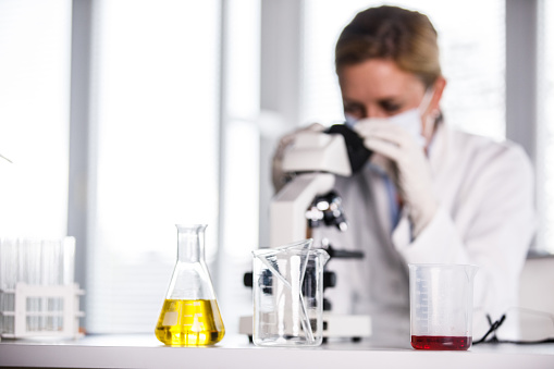 Copy space shot of diligent mid adult female scientist looking through the microscope and analyzing a virus sample for research. She is wearing protective gloves and surgical mask.