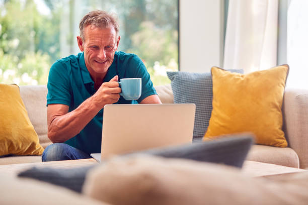 Senior Retired Man Sitting On Sofa At Home Shopping Or Booking Holiday On Laptop stock photo
